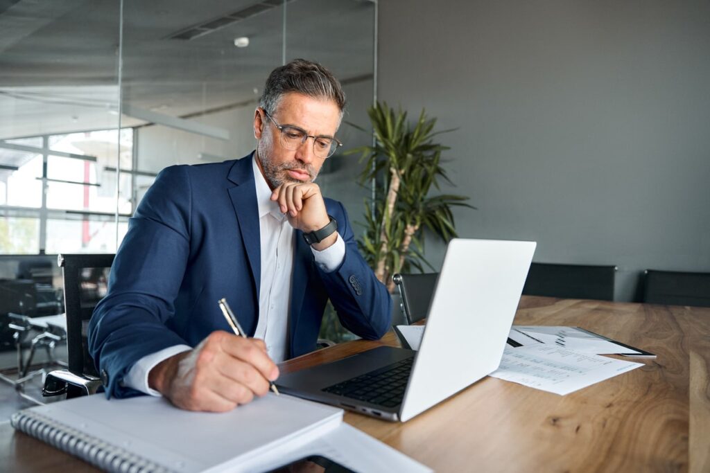 business man executive sitting at desk working on laptop writing notes