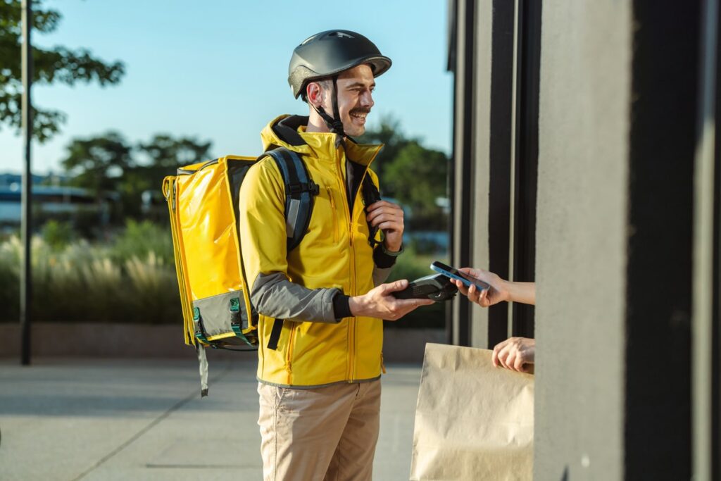 A delivery courier wearing a yellow jacket and helmet hands a payment device to a customer at the doorstep