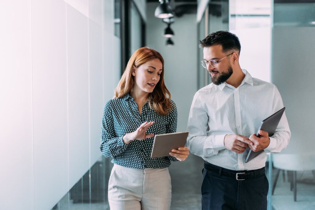 Two finance professionals reviewing data on a tablet while walking through a modern office hallway.