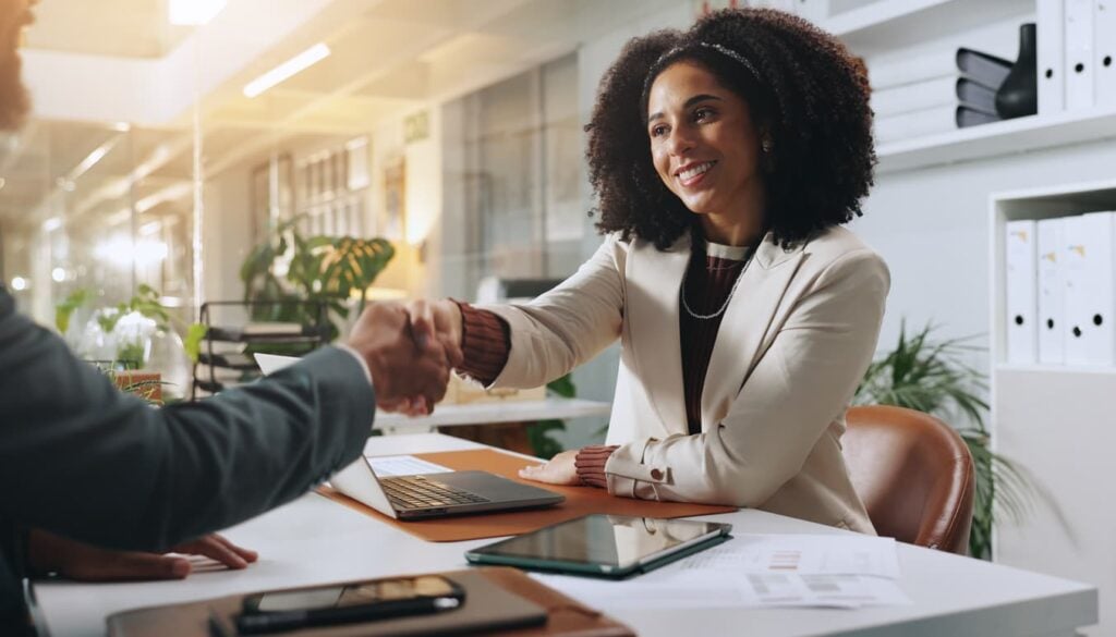 Hiring manager shaking hands with a job candidate across a desk in a modern office setting