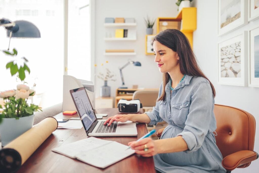 Woman in a denim shirt working at a wooden desk, reviewing data on a laptop and taking notes in a notebook, in a bright home office.