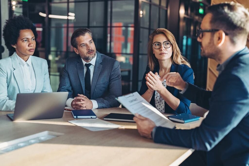 Four finance executives in business attire discuss a document during a leadership meeting at a conference table.