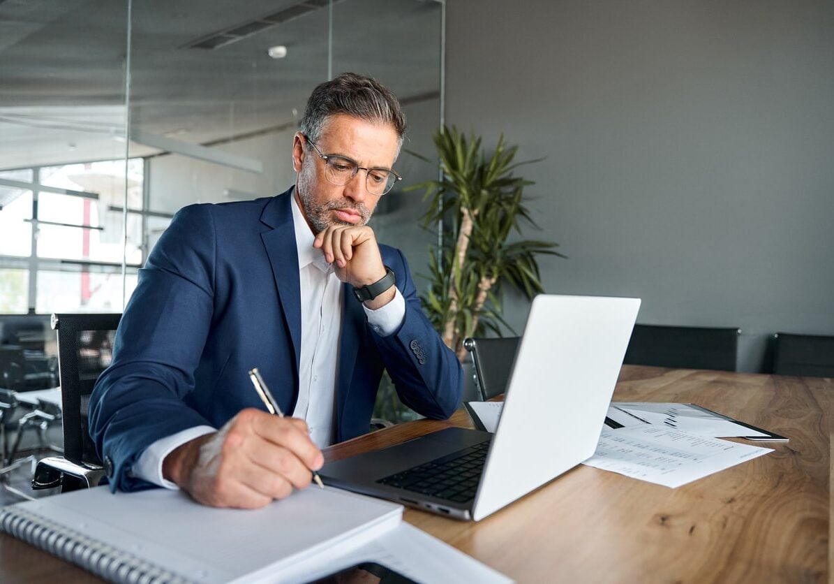 business man executive sitting at desk working on laptop writing notes
