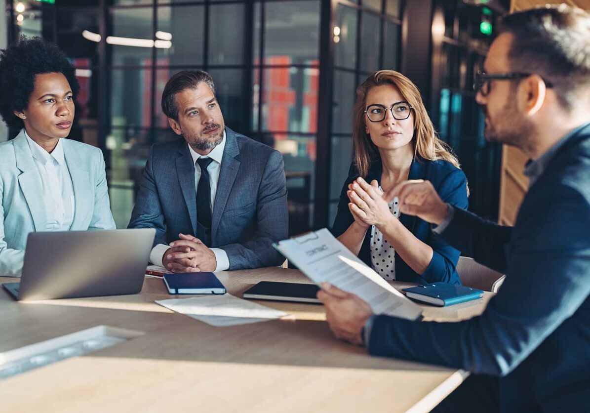 Four finance executives in business attire discuss a document during a leadership meeting at a conference table.