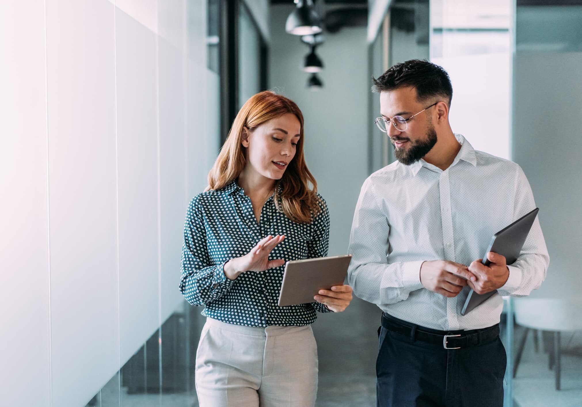 Two finance professionals reviewing data on a tablet while walking through a modern office hallway.