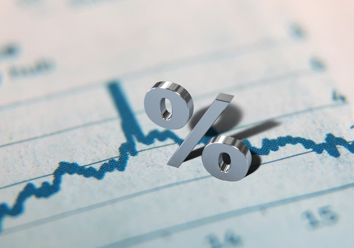 A 3D percent sign casts a shadow over a rising line chart on a financial document, representing interest rate trends.