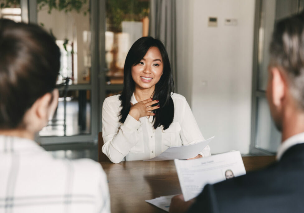 Business, career and placement concept - young asian woman smiling and holding resume while sitting in front of mentor or managers during job interview