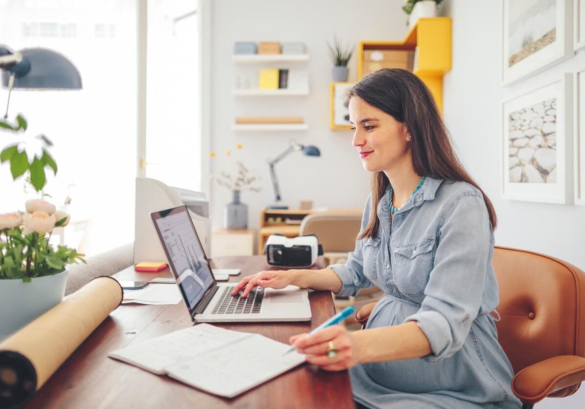 Woman in a denim shirt working at a wooden desk, reviewing data on a laptop and taking notes in a notebook, in a bright home office.
