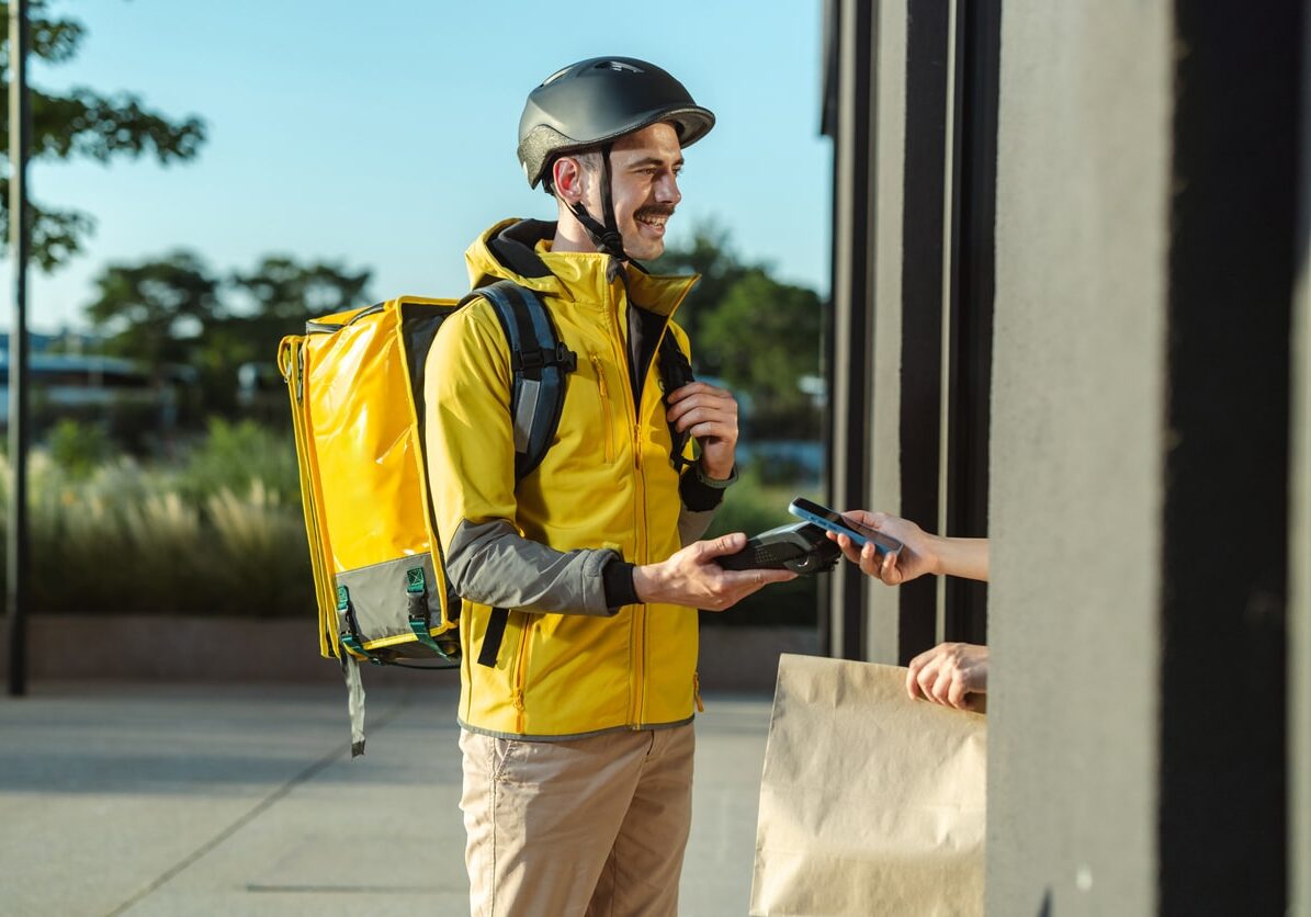 A delivery courier wearing a yellow jacket and helmet hands a payment device to a customer at the doorstep